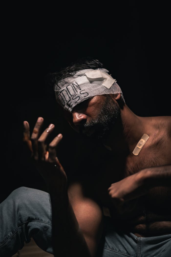 A shirtless man with head and arm bandages, sitting in a pose of contemplation and pain.