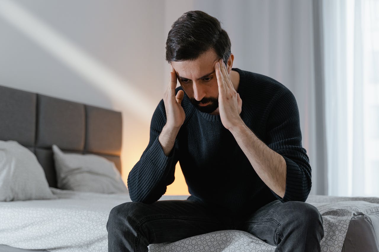 A man in a black sweater sits on a bed, expressing frustration in a bedroom.